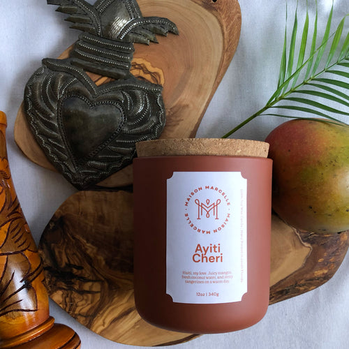 Dark orange Maison Marcelle candle vessel surrounded by Haitian wood vase, Haitian metal art heart, small palm frond, and a mango.